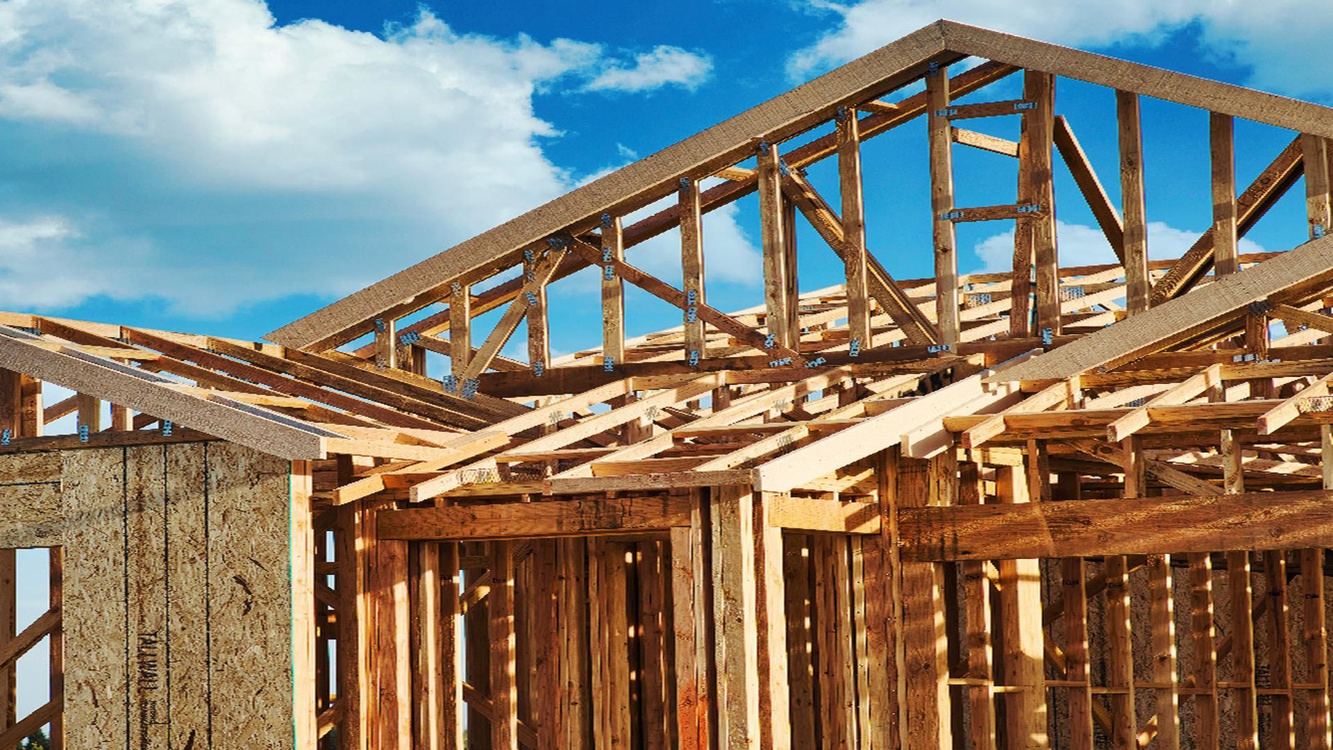 Framing of a single wide manufactured home under construction, showcasing the wooden structure and roof beams against a bright, cloudy sky